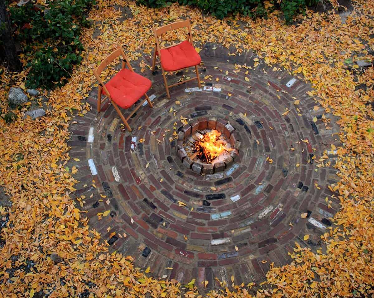 Circular brick fire pit patio with concentric rings of reclaimed bricks surrounding a lit fire, autumn leaves scattered around it