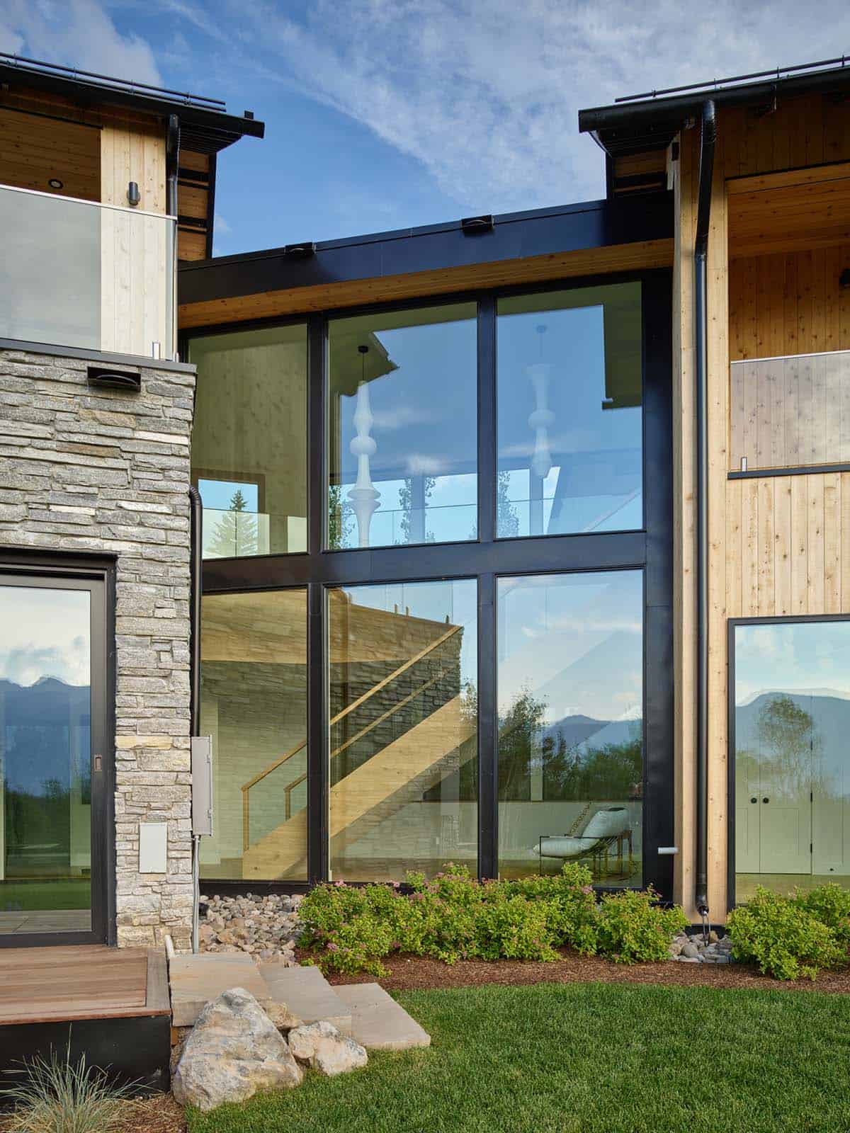 Two-story glass wall reflecting mountain views between stone and wood siding