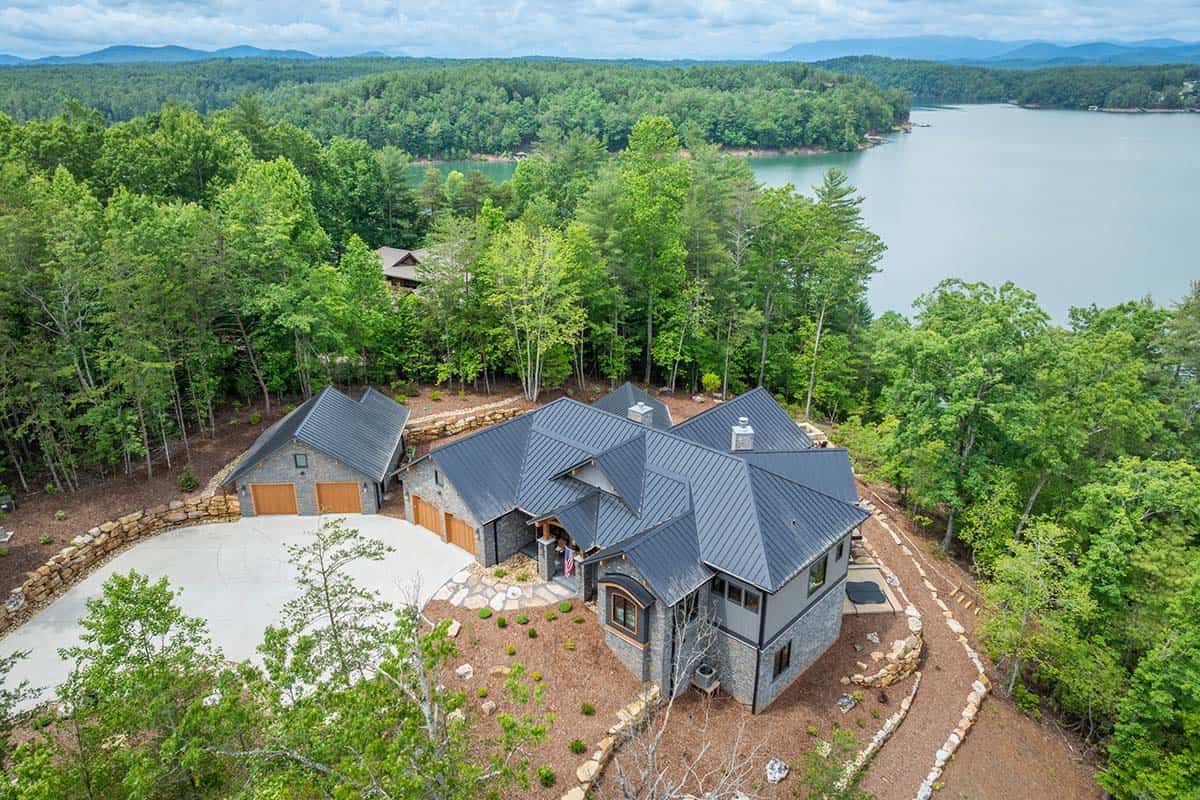 Aerial view of stone lake house with dark standing-seam roof nestled among trees near the water