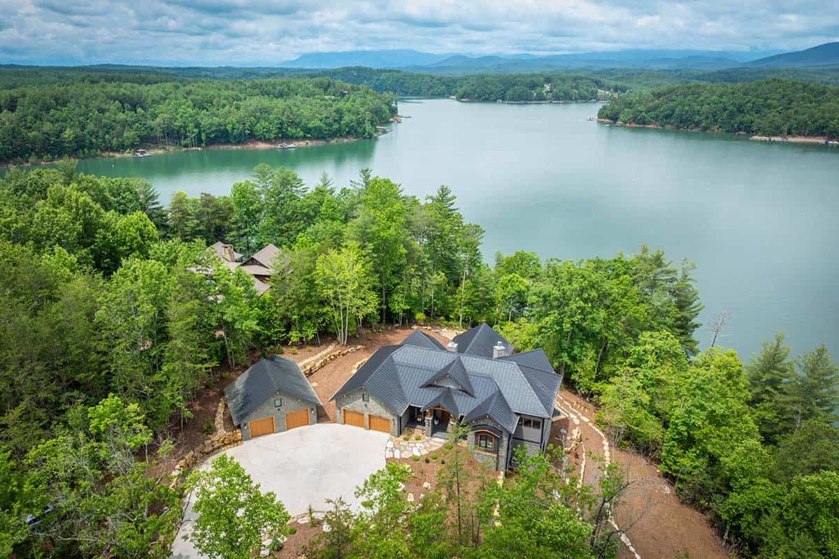 Aerial view of multi-gabled stone house on wooded lake point with mountains in distance