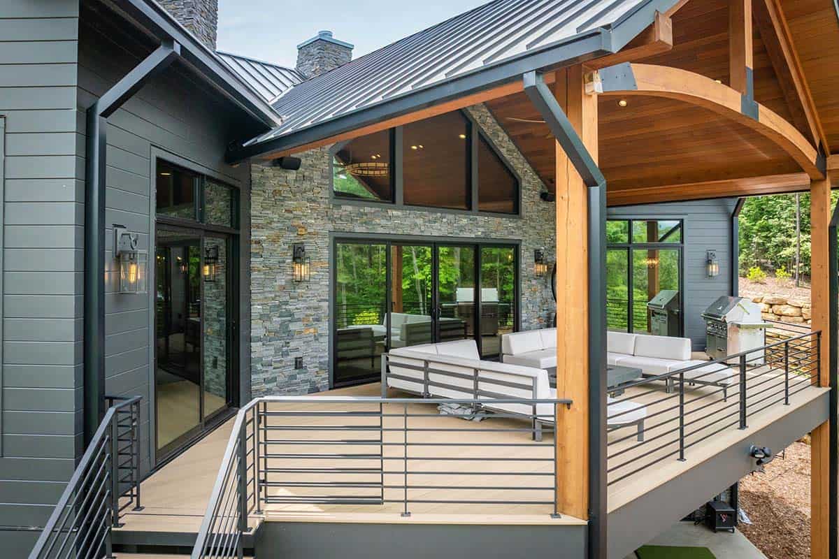 Covered deck with timber trusses, stone wall, and white outdoor furniture overlooking the forest