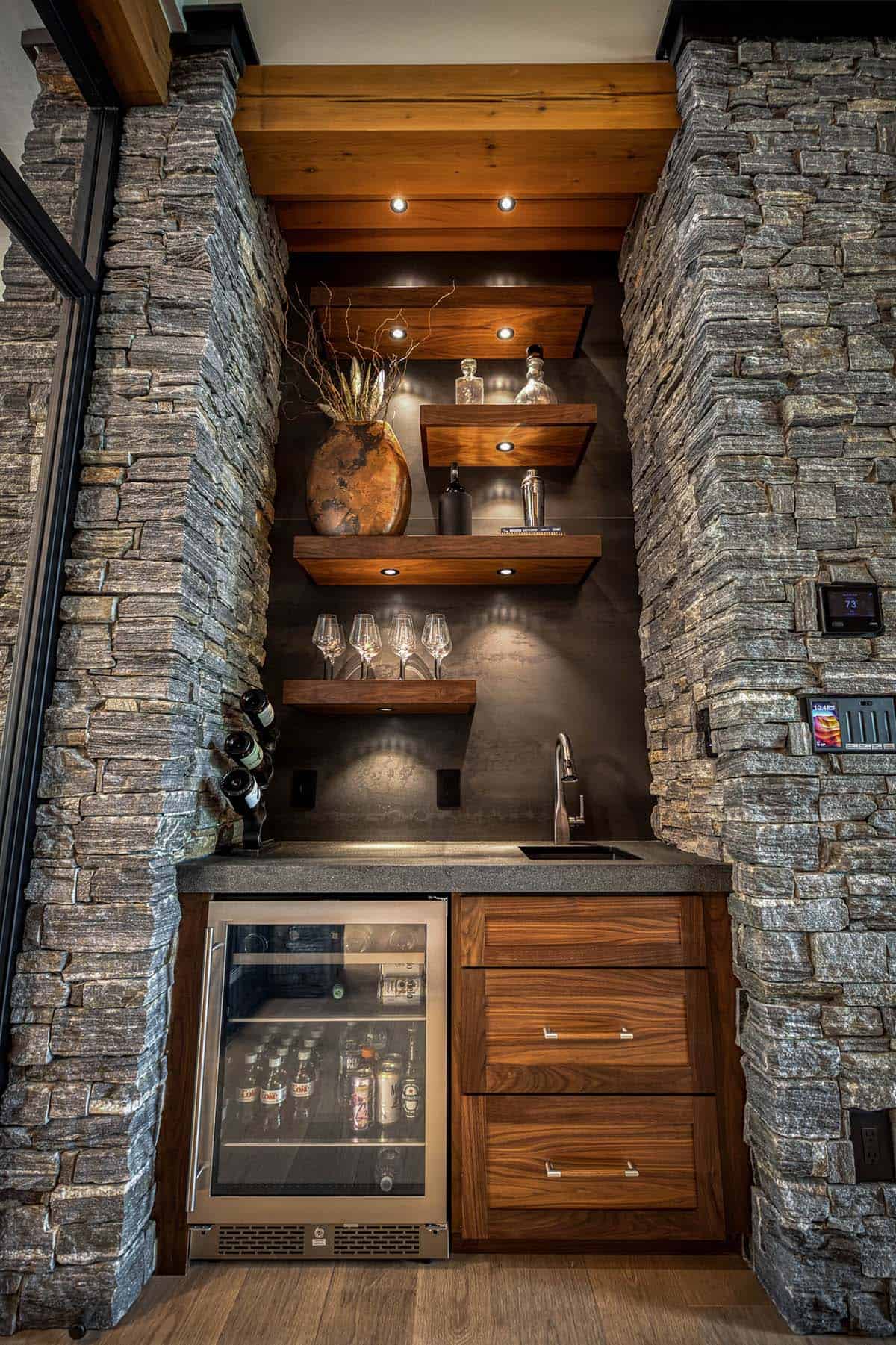 Full view of stone-framed wet bar with floating shelves, sink, and beverage fridge