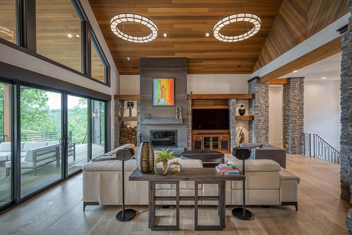 Great room with wood ceiling, ring pendants, stone fireplace, sectional sofa, and deck view