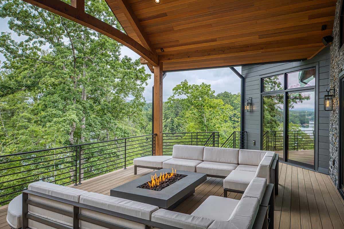 Deck seating area under timber roof with sectional sofa, fire table, and tree-lined view