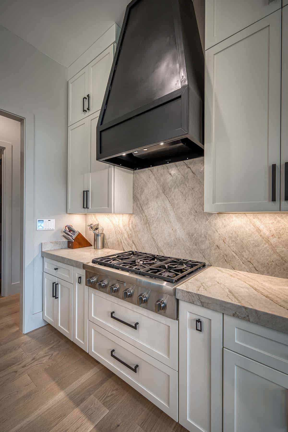 Close-up of white kitchen cabinetry with black range hood, gas cooktop, and stone backsplash