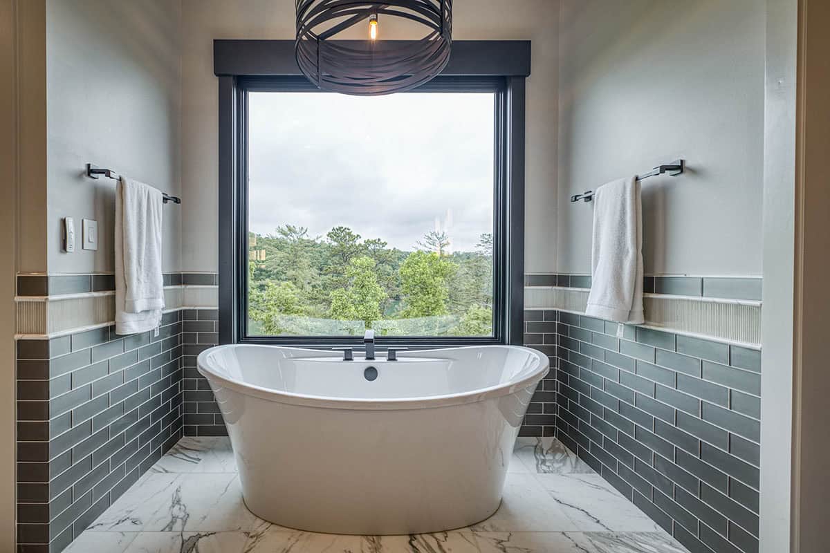 Freestanding soaking tub framed by subway tile and window with wooded view