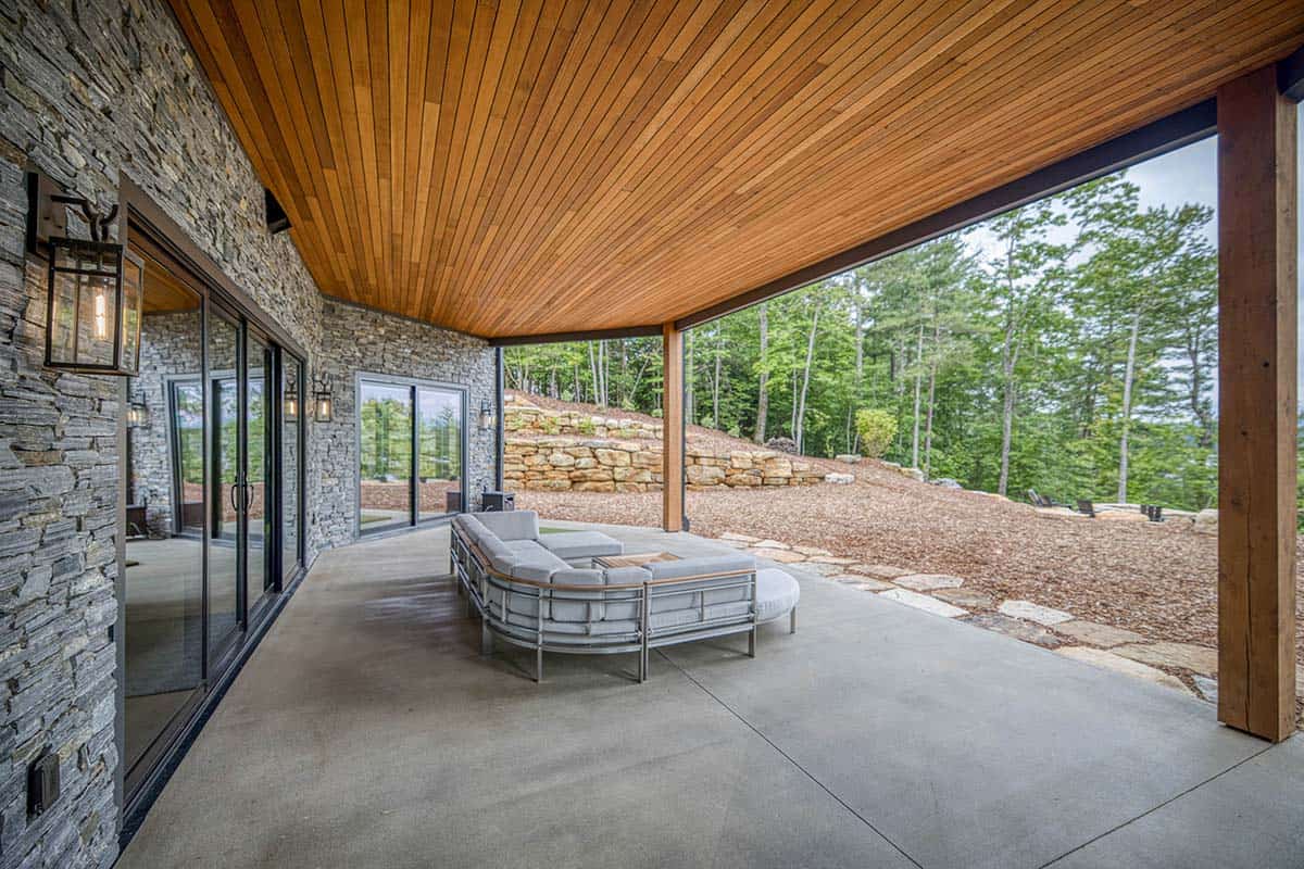 Covered patio with wood ceiling, stone wall, curved outdoor sofa, and wooded hillside view