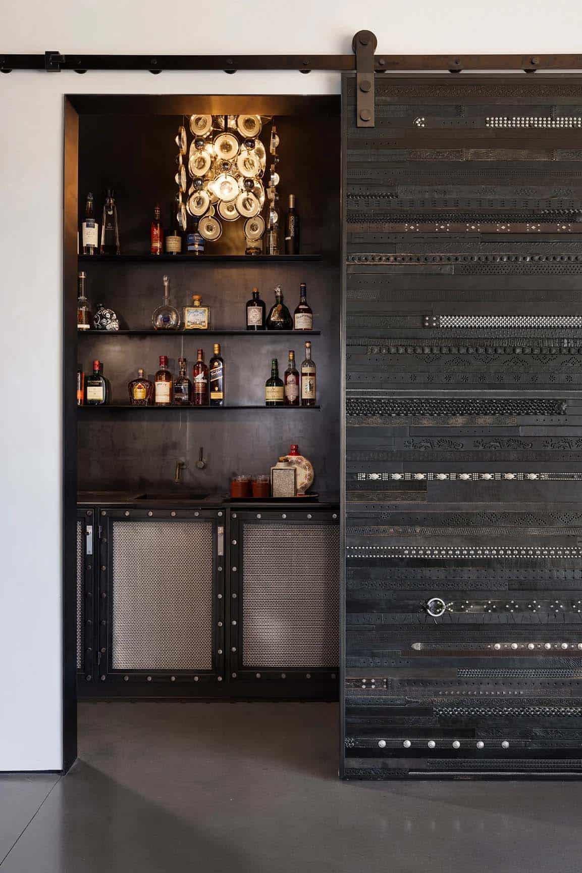 Wet bar nook with belt-clad sliding door, brass chandelier, and shelved liquor bottles