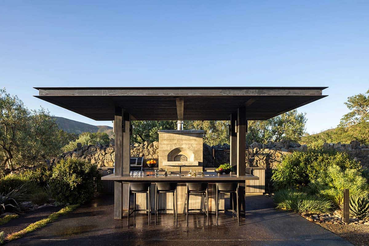 Outdoor kitchen pavilion with wood-fired pizza oven, bar seating, and steel roof canopy