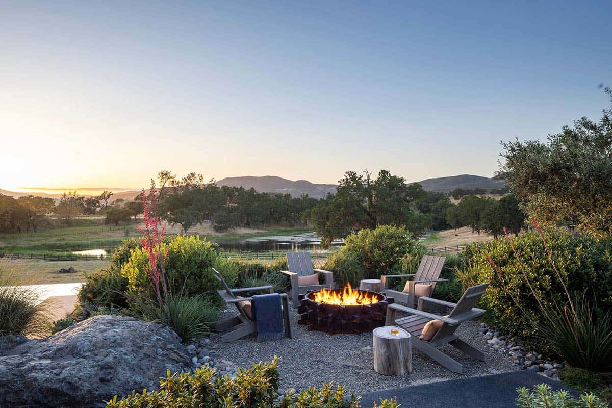 Fire pit surrounded by Adirondack chairs overlooking a pond and distant hills at dusk
