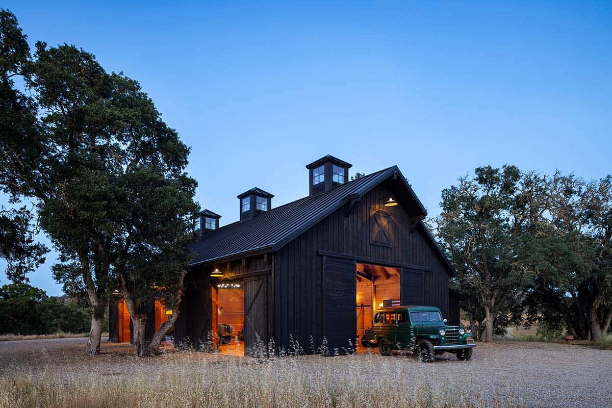 Black board-and-batten barn with glowing interior and vintage truck parked out front at twilight