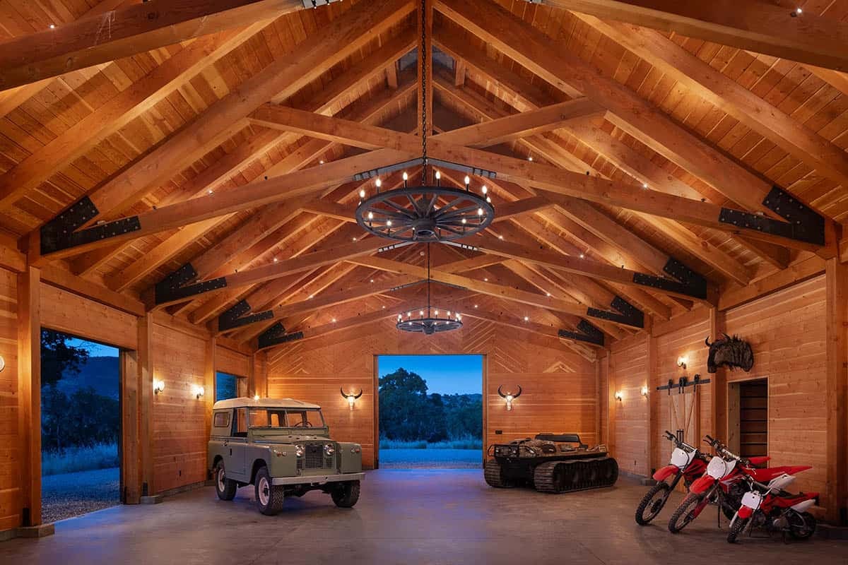 Timber-framed barn interior with exposed trusses, wagon-wheel chandeliers, and vintage vehicles