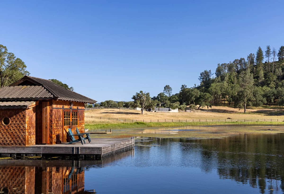 Shingle-clad boathouse on a still pond, with Adirondack chairs on its dock