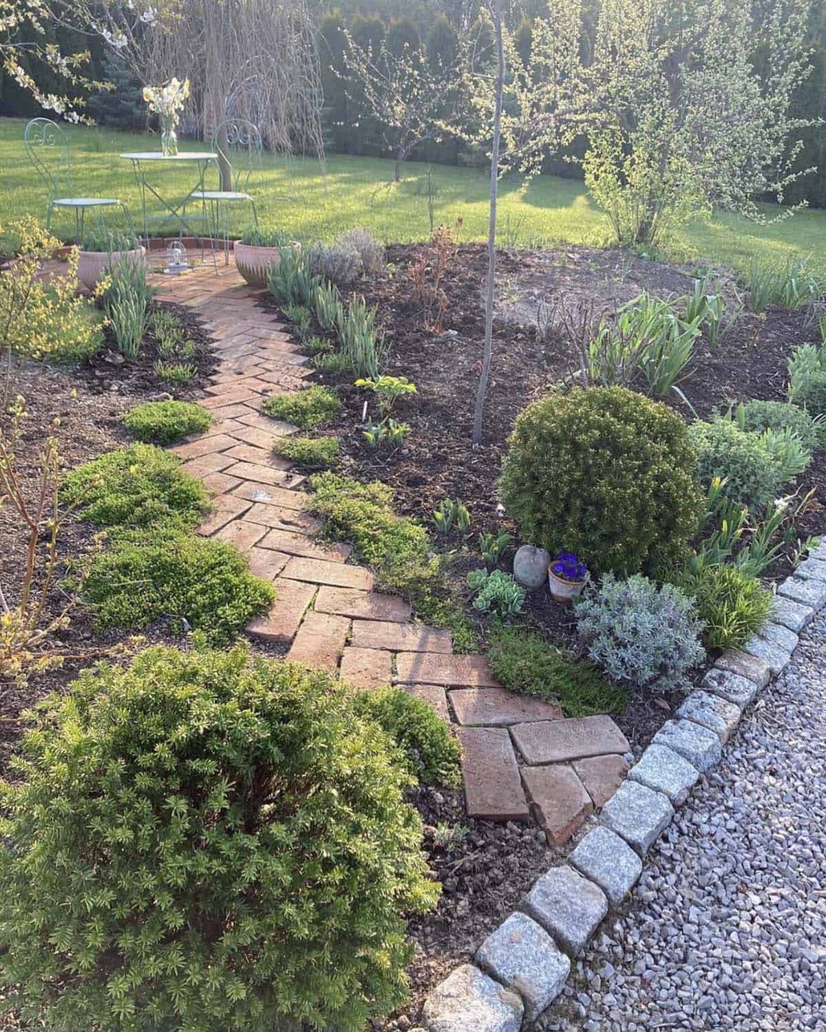 Curving brick path winding through a planted garden bed toward a small bistro table and chairs