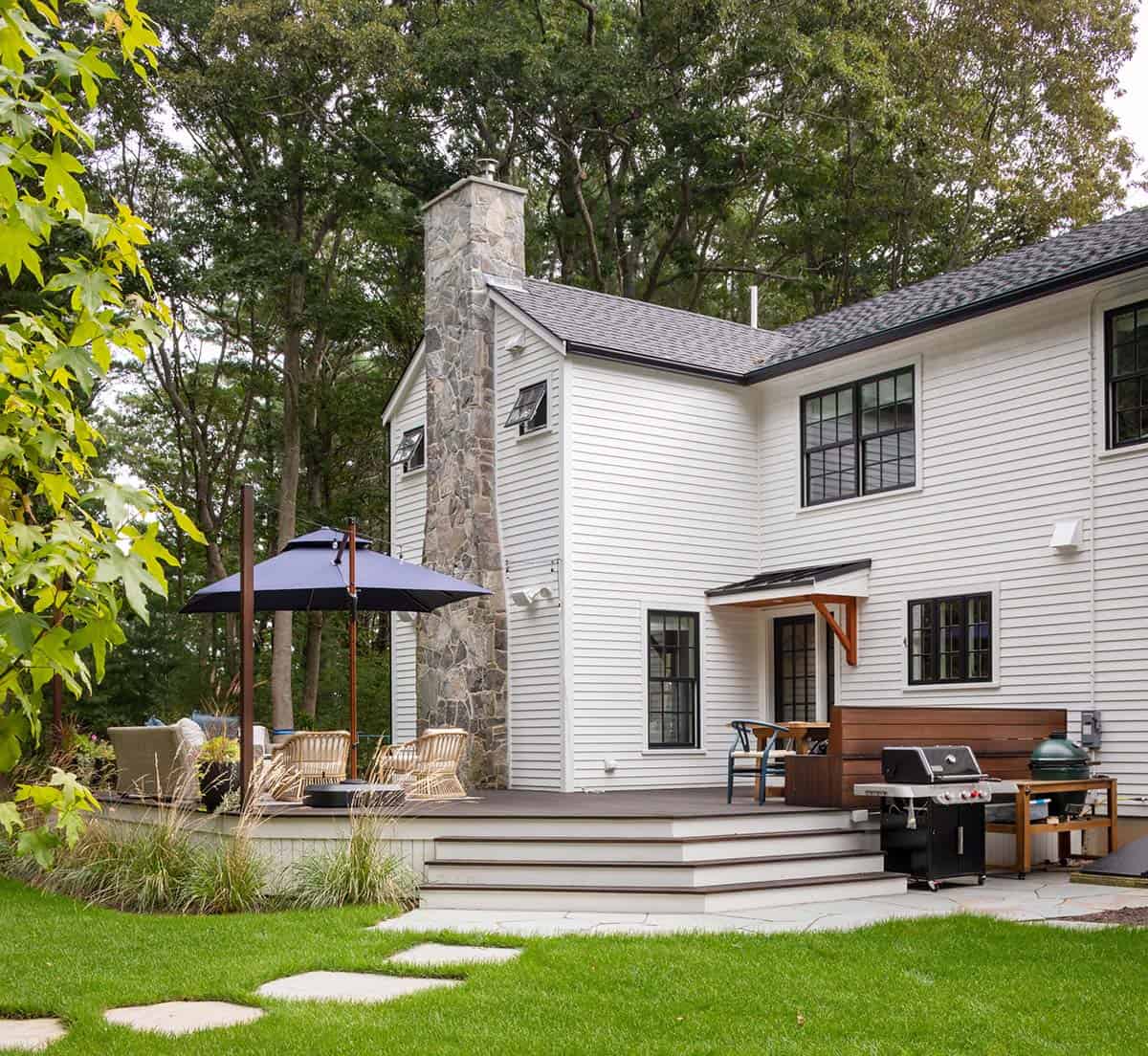 Wood deck with grill station, wicker seating, and navy umbrella beside white farmhouse exterior
