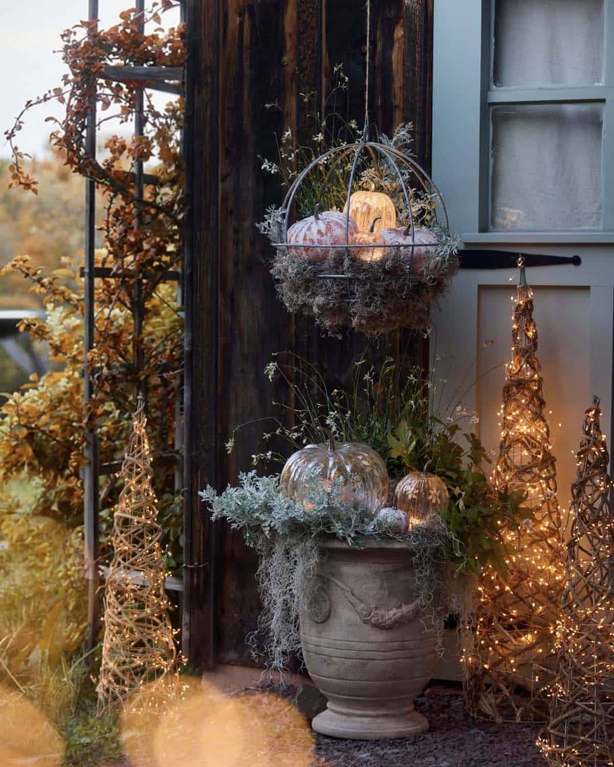 Hanging pumpkin basket beside a planter and lit twig trees at dusk
