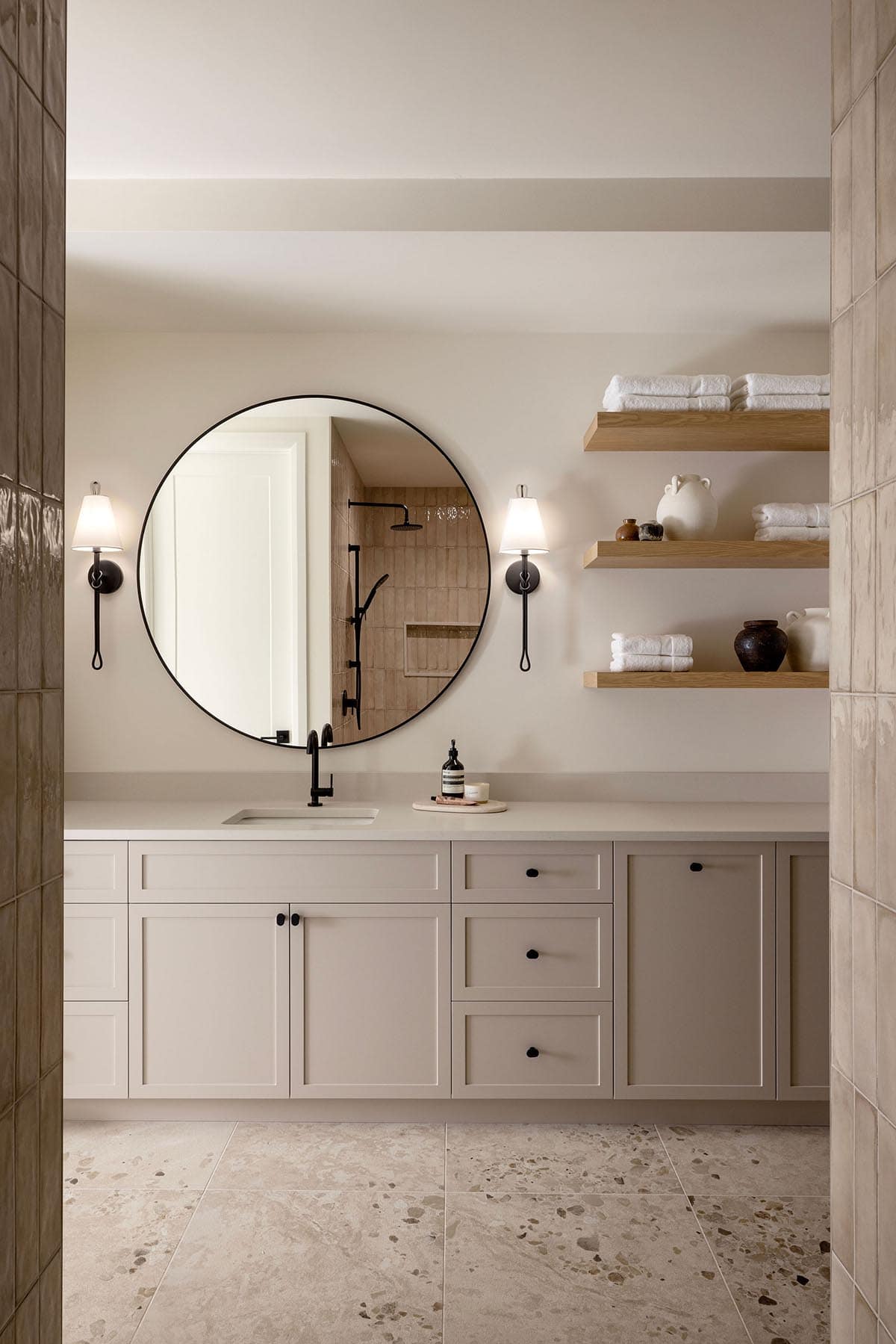Bathroom vanity with round black-framed mirror, sconces, and floating wood shelves