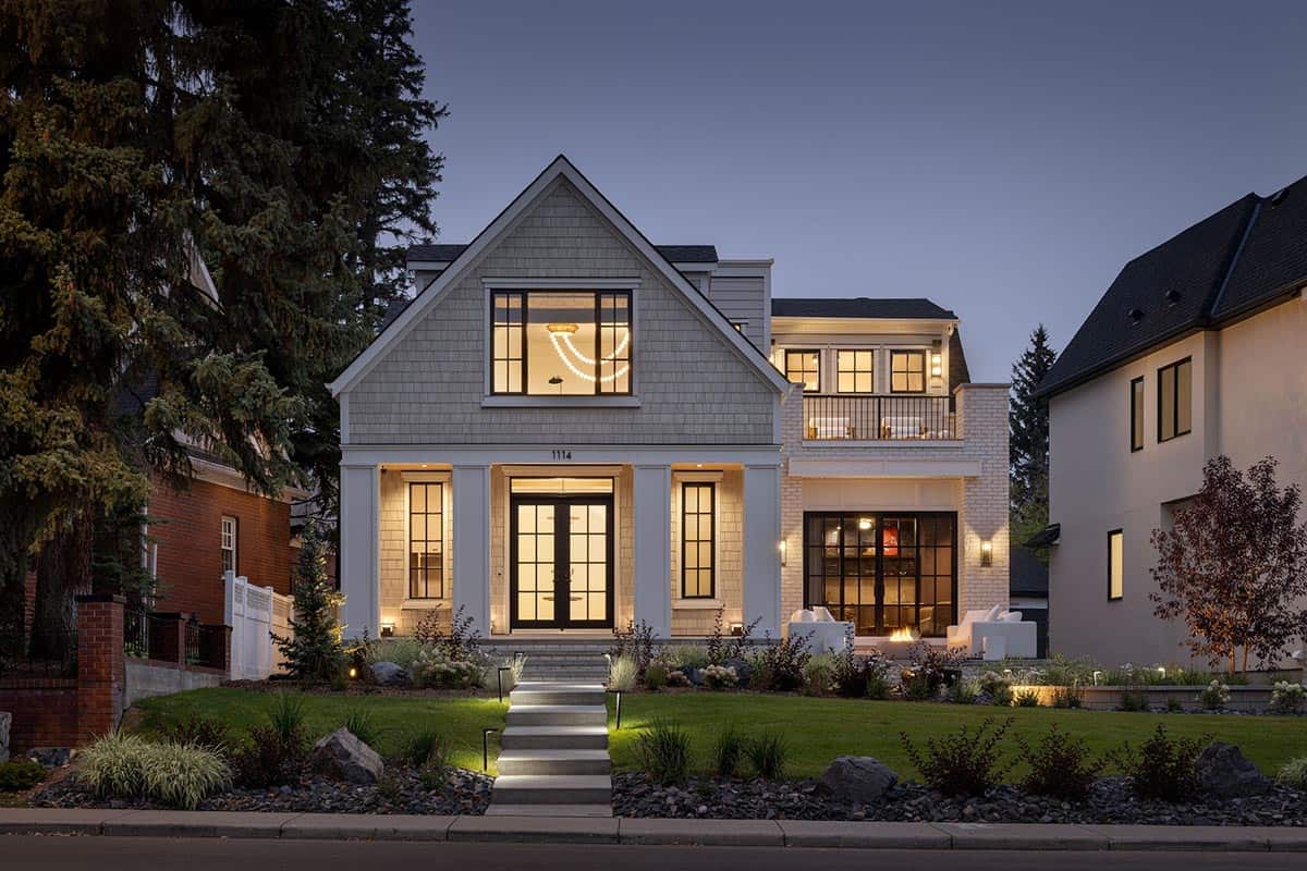 Twilight exterior of Hamptons-style estate with illuminated windows and landscaped front yard