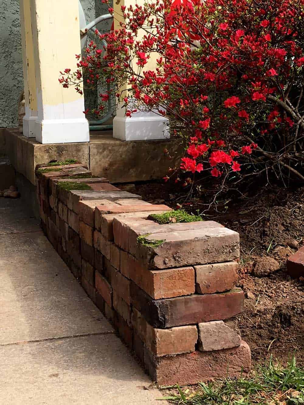 A low, uneven reclaimed brick retaining wall stacked in a higgledy-piggledy style beside a front porch, with a red azalea blooming behind it