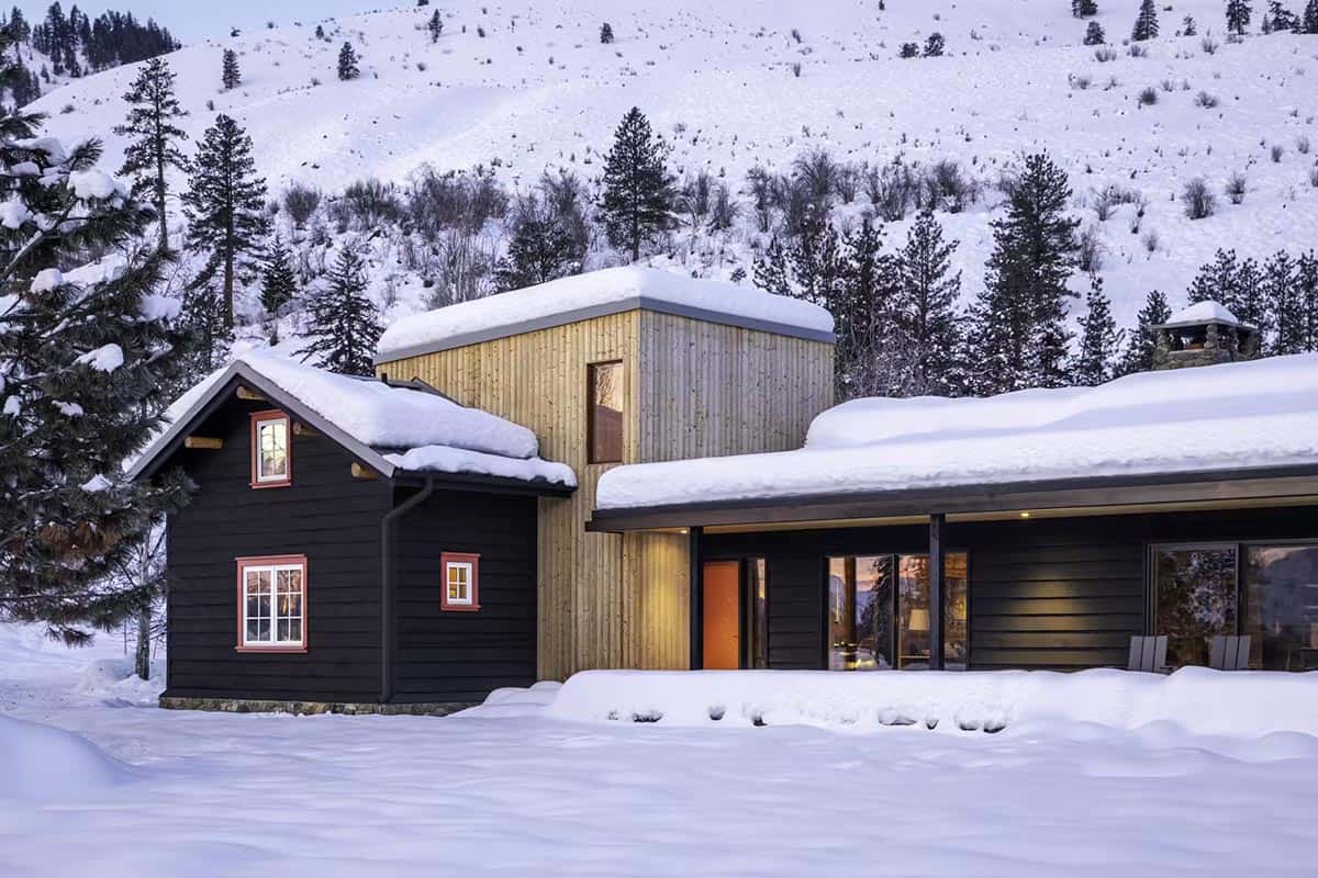 Exterior view of black cabin with light wood addition and red door, snow-covered roof