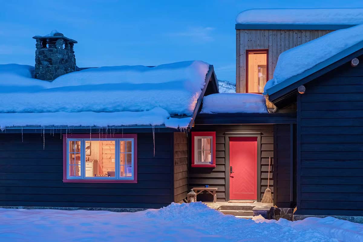 Twilight view of black cabin with red door, glowing windows, and stone chimney