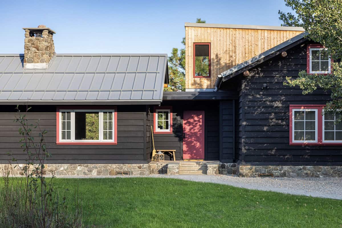 Black cabin exterior with red-trimmed windows and wood addition against blue sky