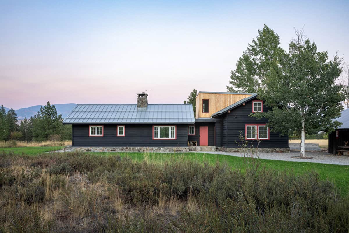 Black cabin with red trim and wood addition at sunset, surrounded by trees