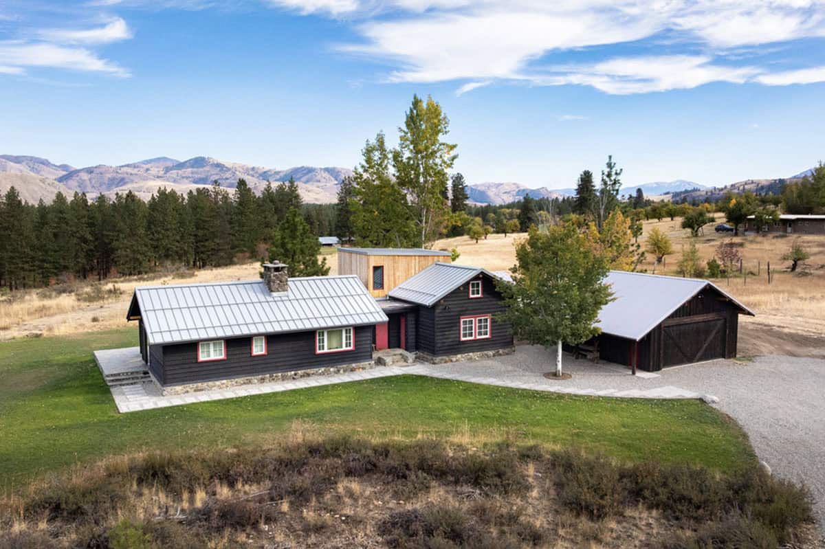 Aerial view of black cabin complex with barn, mountains, and open lawn