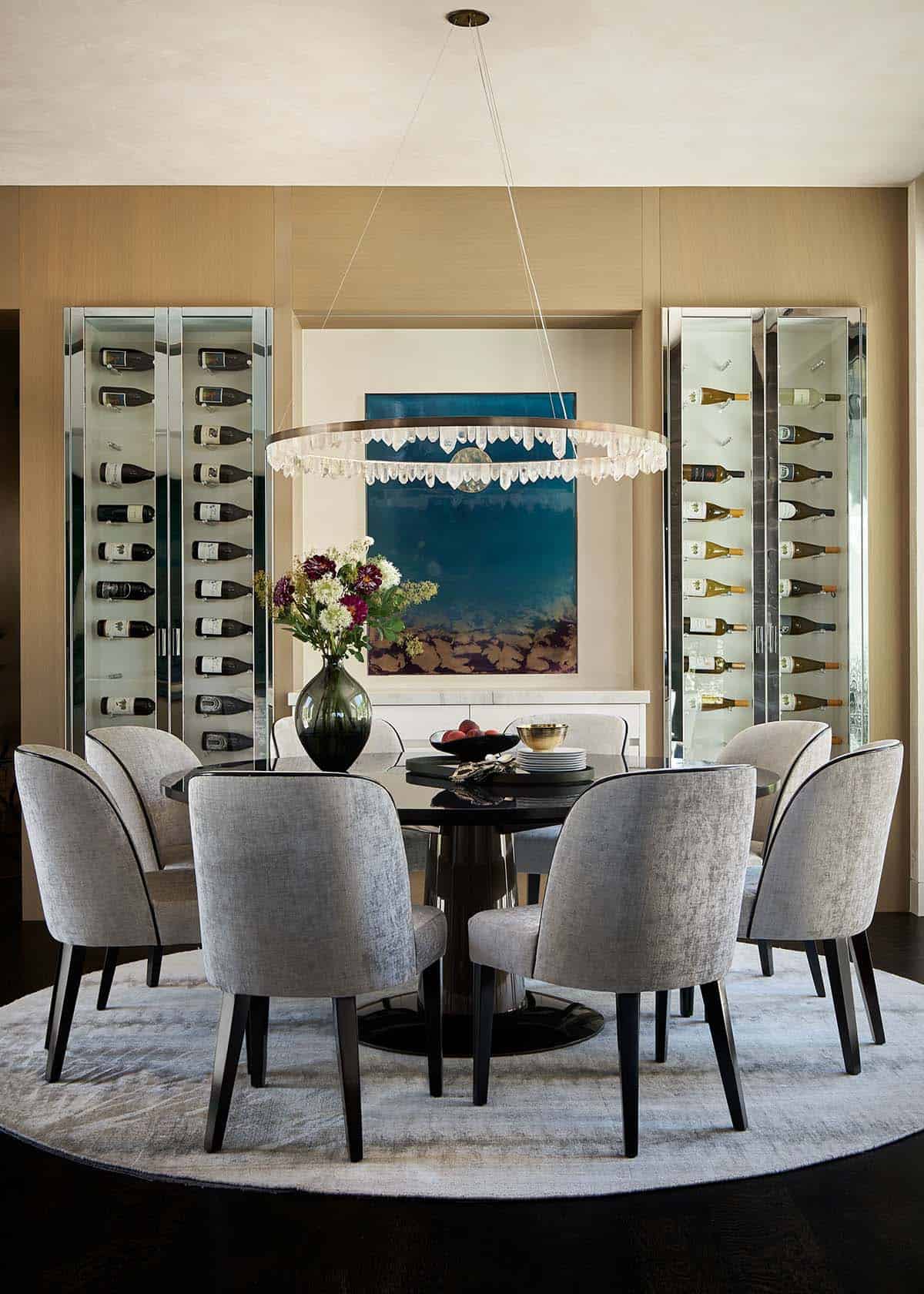 Round dining table with gray upholstered chairs beneath a crystal ring chandelier, flanked by glass wine cabinets