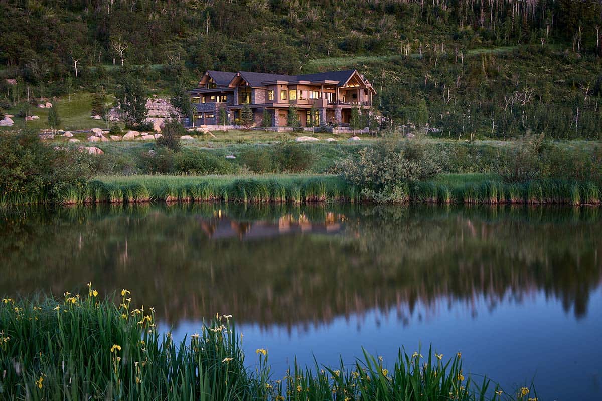 Mountain home nestled in forested hillside, lit windows reflected in pond
