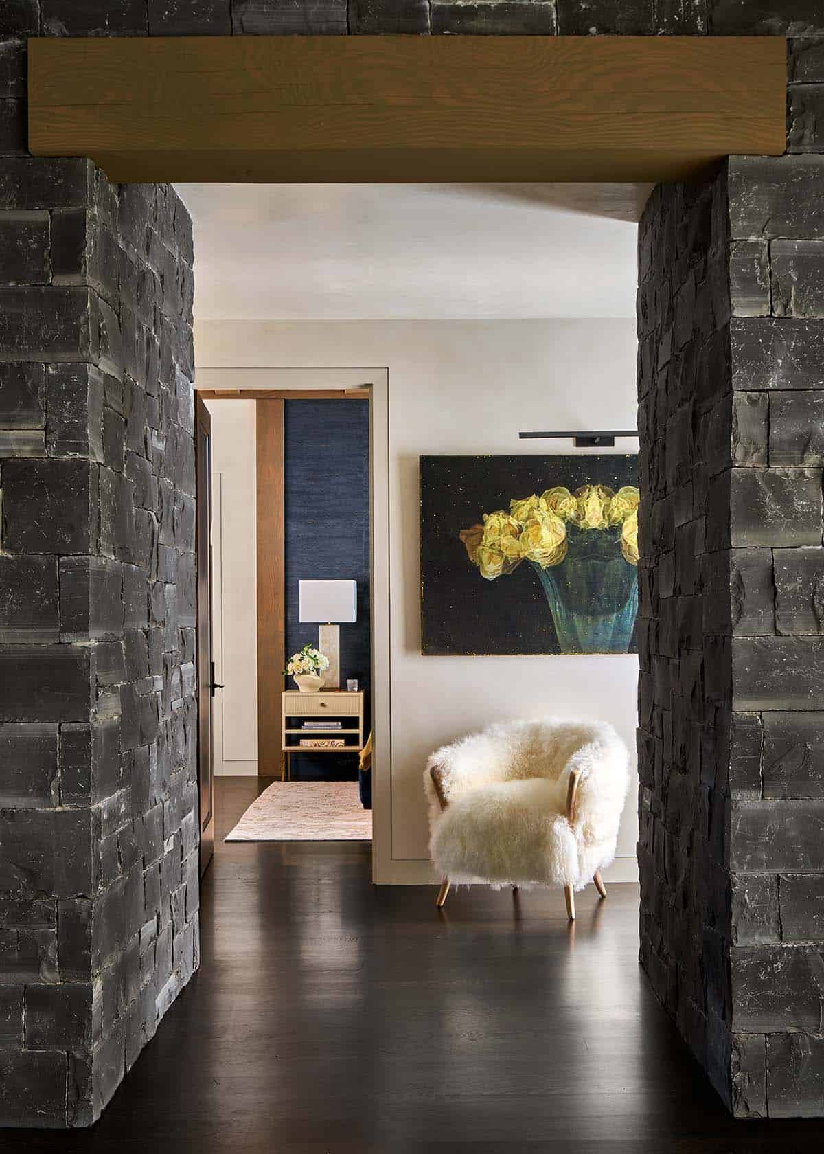 Hallway between stacked-stone walls leading to bedroom with floral artwork and shearling chair