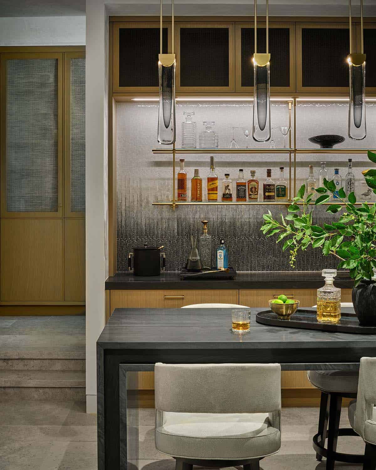 Wet bar with brass pendant lights, glass shelving, and liquor bottle display