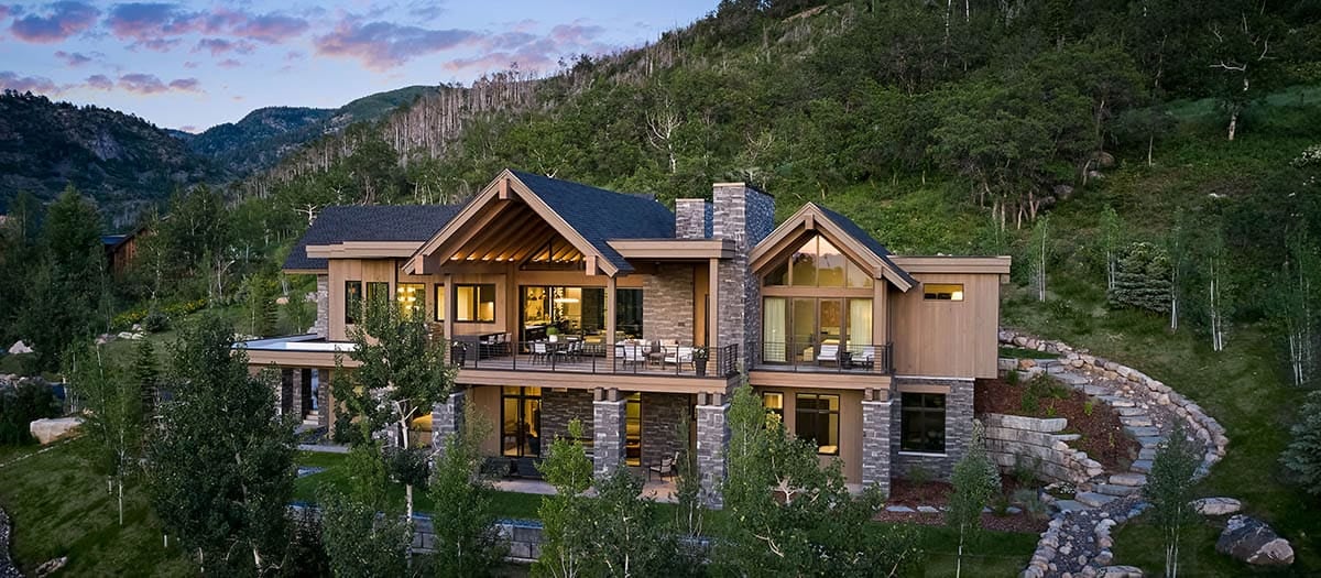 Wide aerial view of stone-and-timber mountain home surrounded by trees at dusk