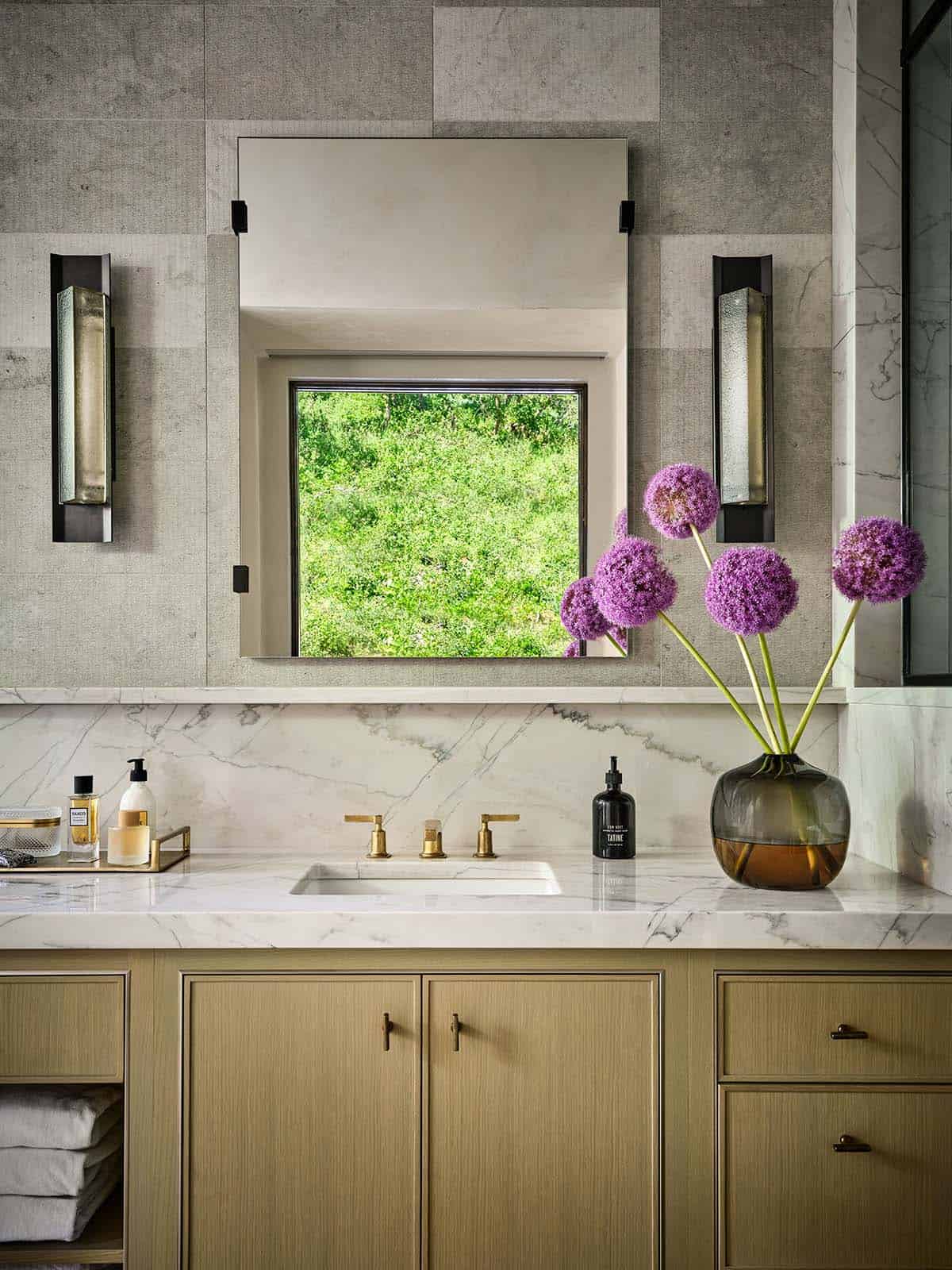Bathroom vanity with marble backsplash, brass faucet, and purple allium in a vase
