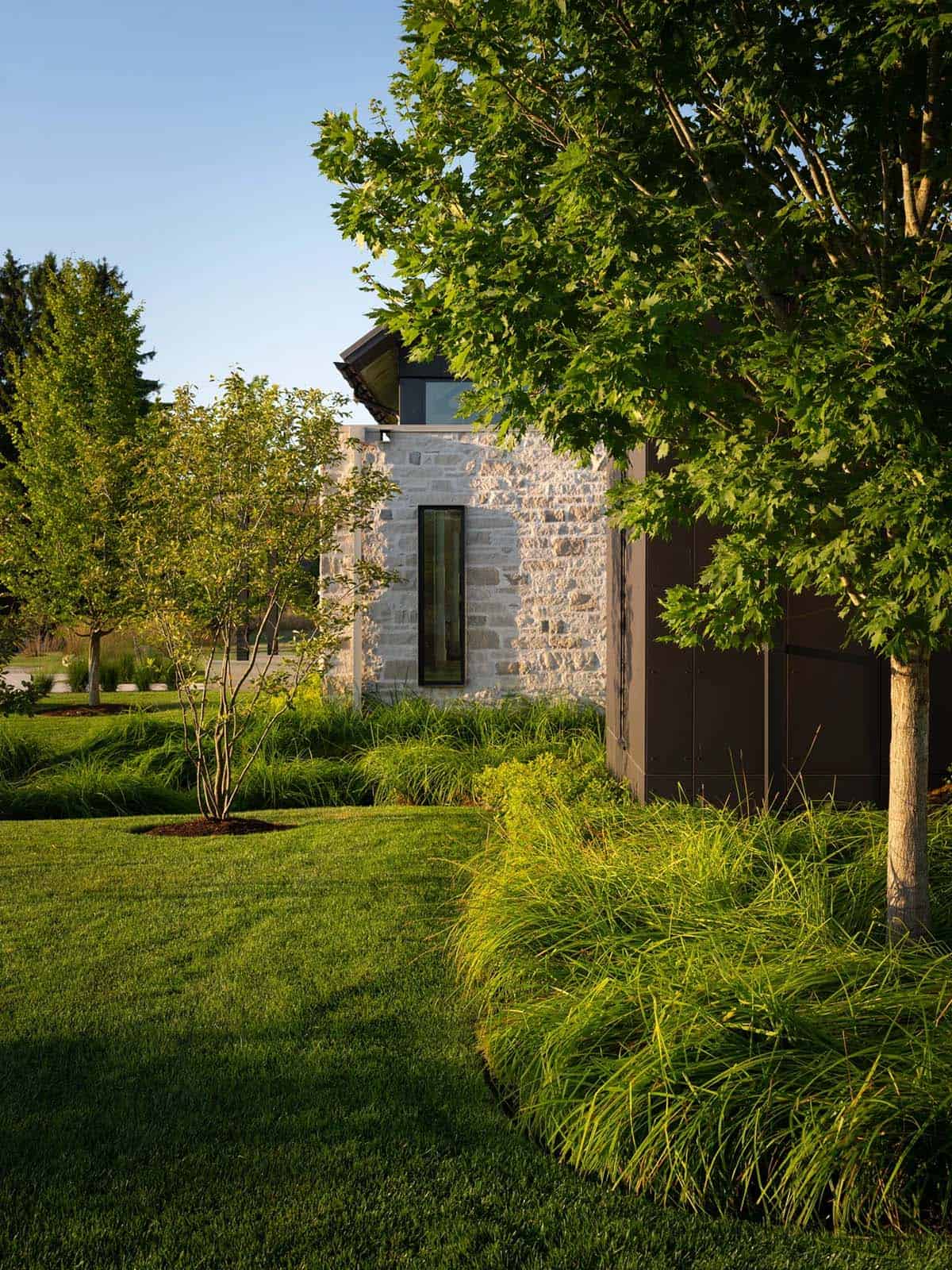 Stone corner of home with narrow window, framed by maple trees and ornamental grasses
