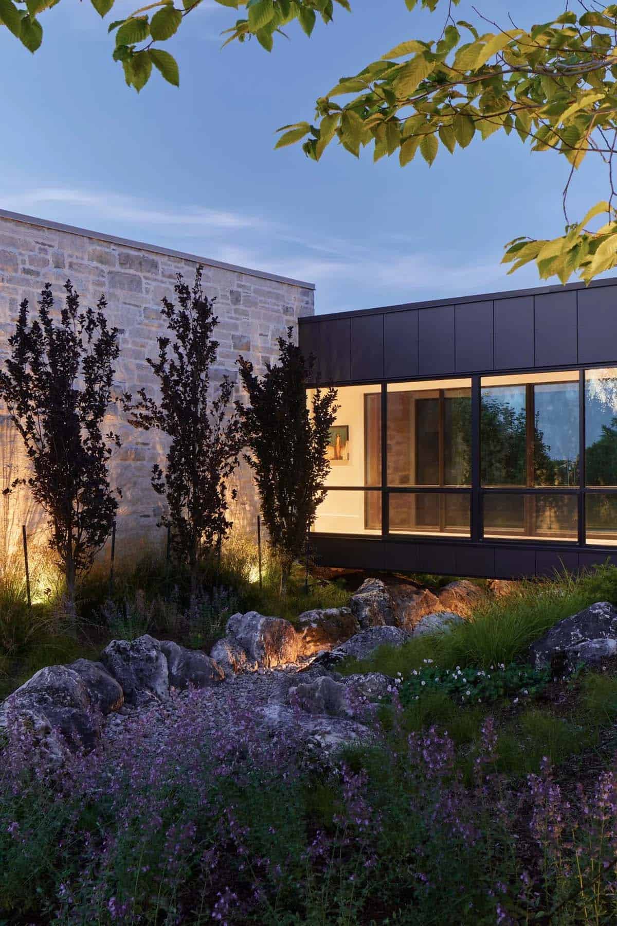 Stone and black-paneled wing connected by glass walkway, lit at dusk