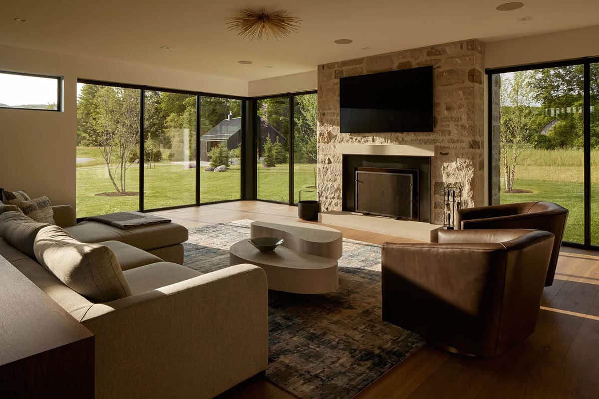 Living room with stone fireplace, leather chairs, and floor-to-ceiling windows overlooking lawn