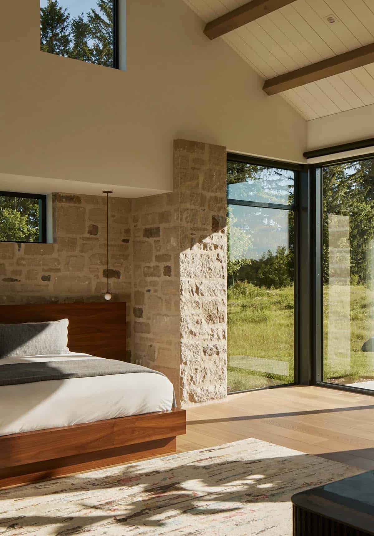 Bedroom with stone accent wall, walnut bed frame, and corner windows to meadow