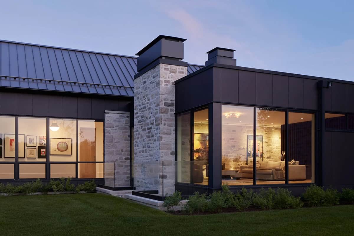 Dark-paneled home exterior at dusk with stone chimney and glowing interior