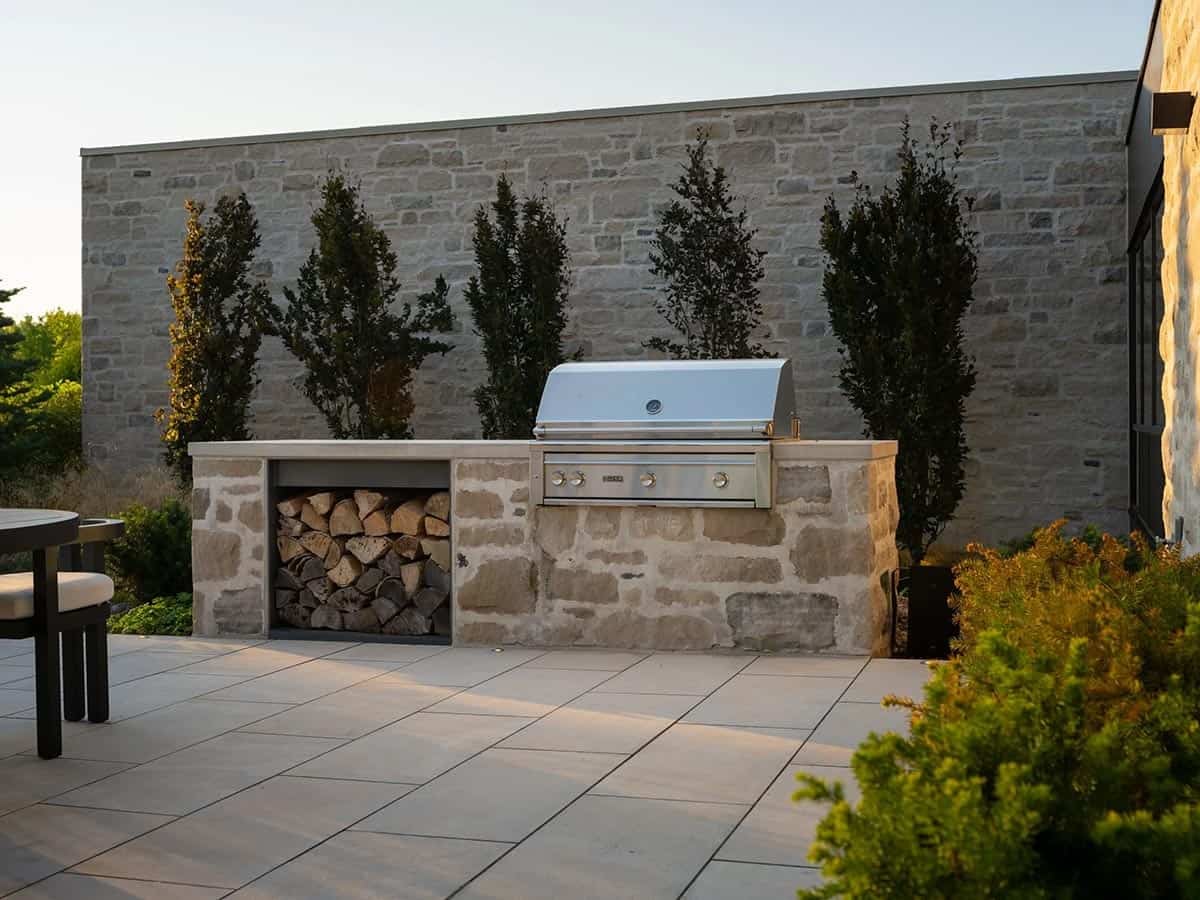 Outdoor kitchen with built-in grill and firewood storage against stone wall