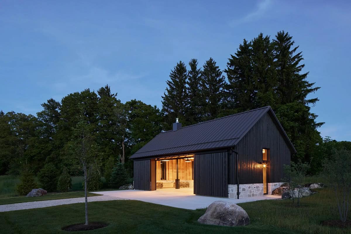 Garage pavilion glowing at dusk with open doors and pine tree backdrop