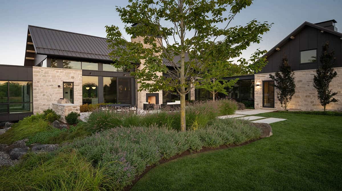 Courtyard view of stone chimney, dark-clad wings, and lush garden beds at dusk