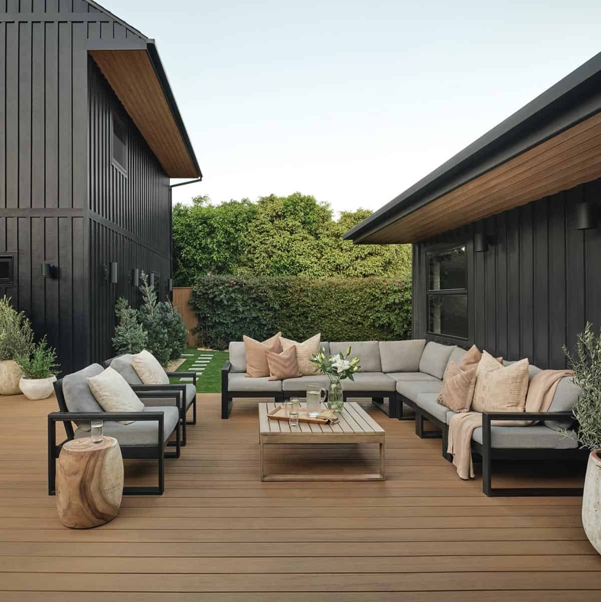 Modern deck with black modular sectional, gray cushions, and blush throw pillows against black barn-style home