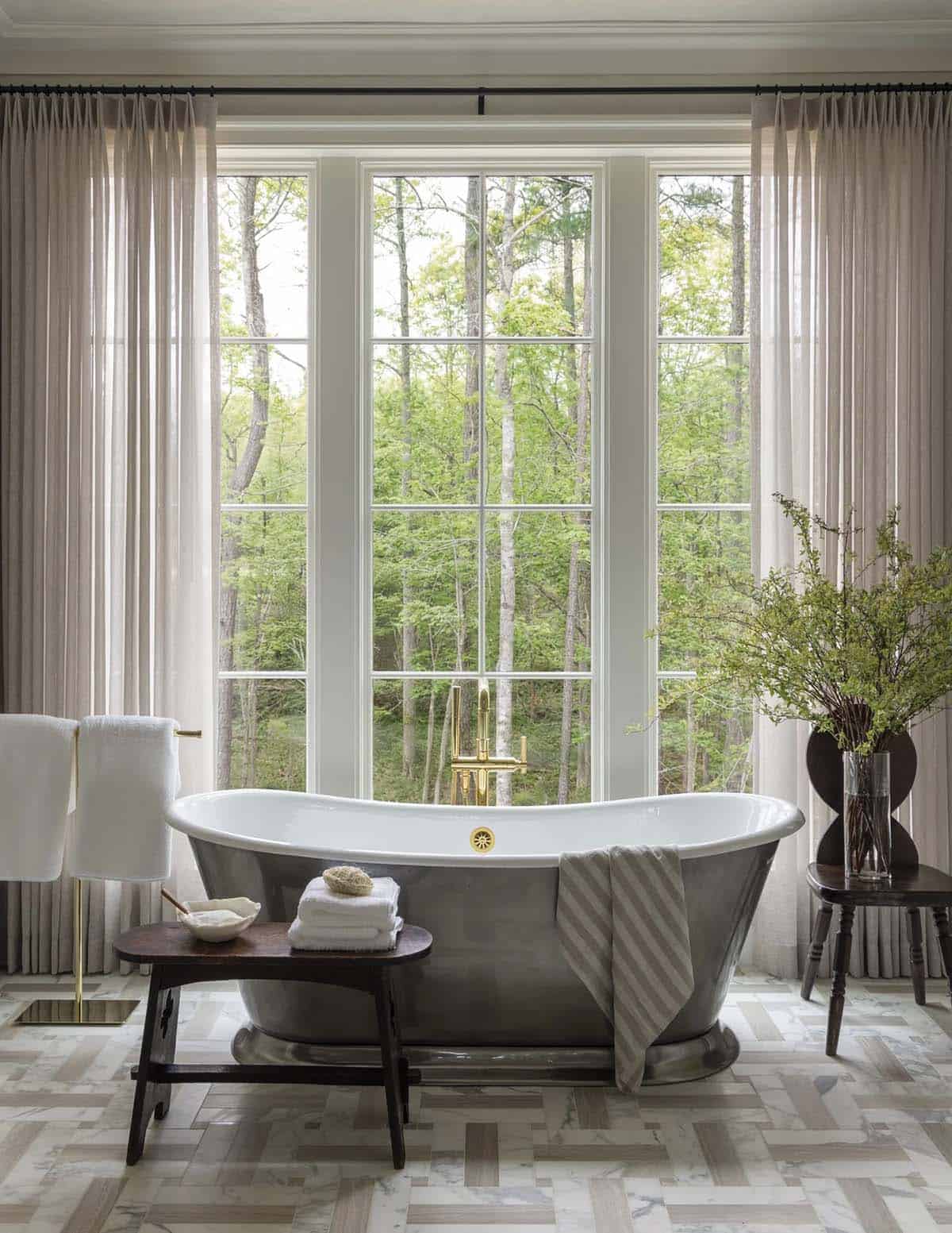 Freestanding metal soaking tub in front of tall windows with brass fixtures