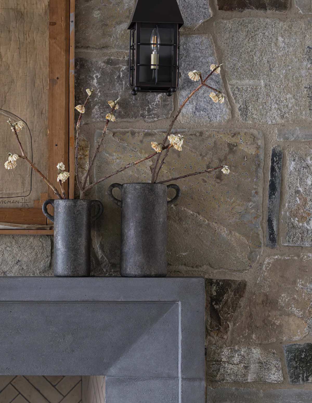 Detail of two textured vases with dried branches on a stone mantel beside a lantern sconce