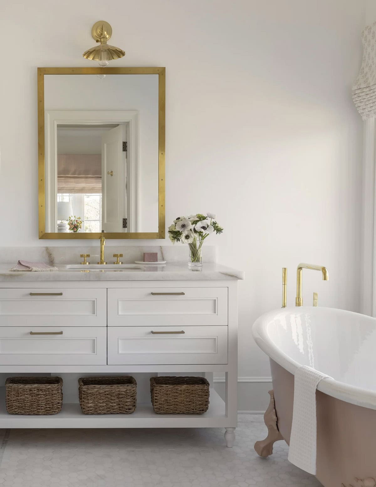 Blush clawfoot tub beside a white vanity with brass mirror and woven baskets