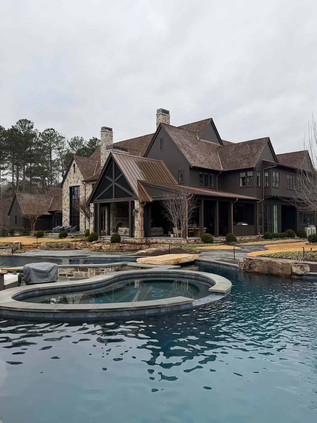 Rear exterior of the dark stone-and-shingle house overlooking the pool on a cloudy day