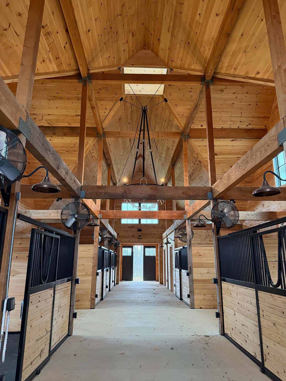 Interior of a wood-beamed horse barn aisle with stalls and hanging fans