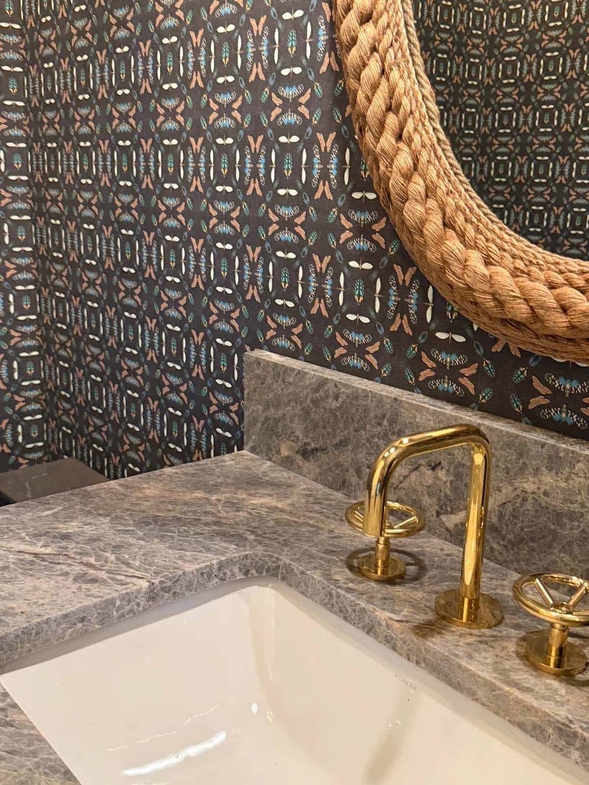 Powder room detail with patterned wallpaper, braided rope mirror, and brass faucet