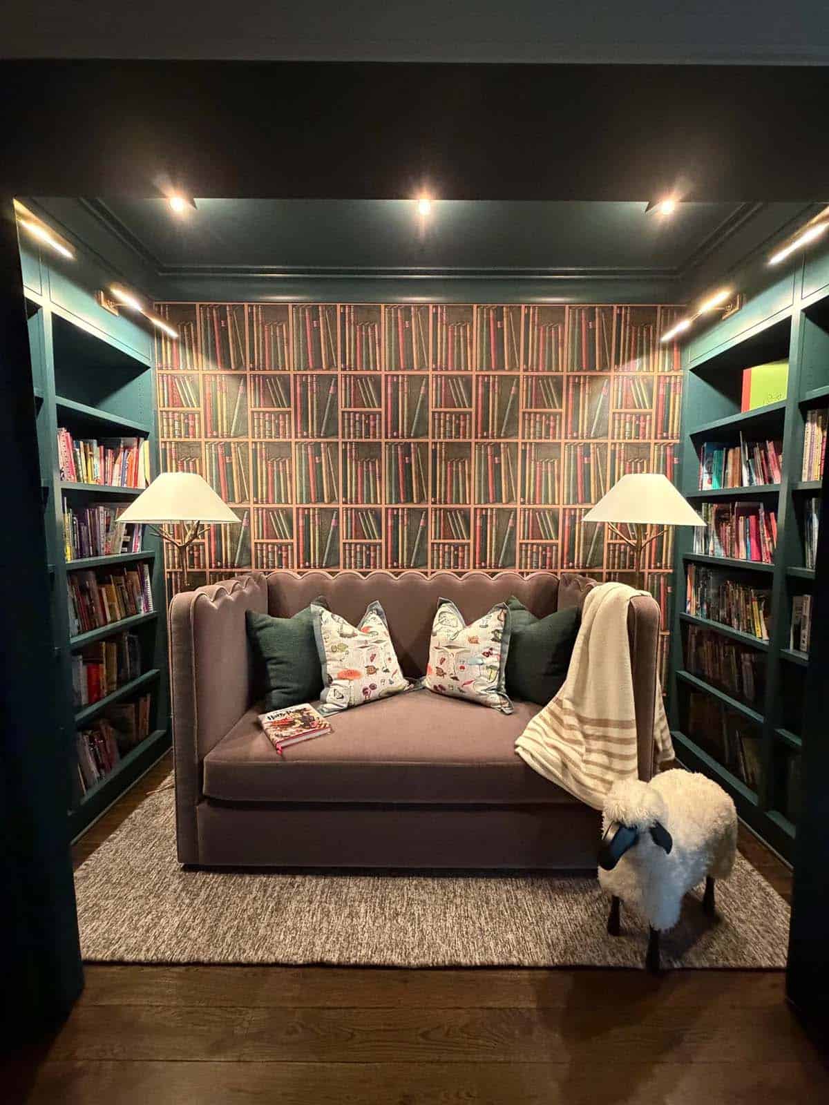 Teal reading nook with bookshelf wallpaper, mauve settee, and sheepskin sheep stool